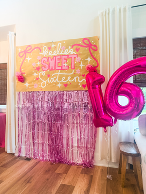 Photo backdrop decorations for a sweet 16 birthday party, including a custom, hand-lettered banner, metallic balloon numbers, and a metallic pink fringe curtain.