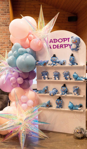 An adopt a derpy party favor station with plushies for a kpop demon hunters party for tweens or kids, next to a diy balloon arch kit.