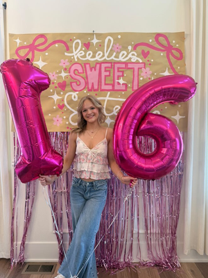 Photo backdrop decorations for a sweet 16 birthday party, including a custom, hand-lettered banner, metallic balloon numbers, and a metallic pink fringe curtain.