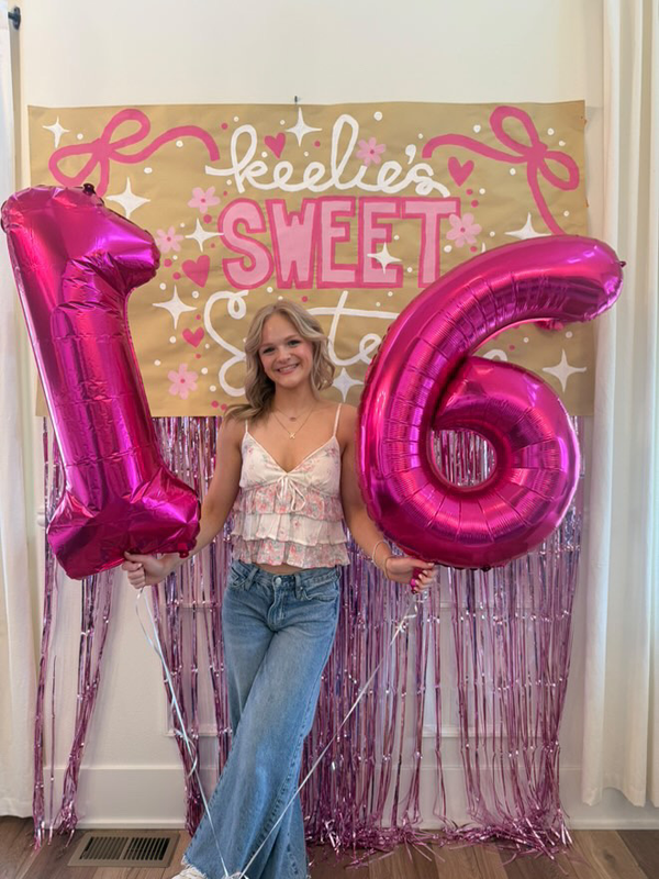 Photo backdrop decorations for a sweet 16 birthday party, including a custom, hand-lettered banner, metallic balloon numbers, and a metallic pink fringe curtain.