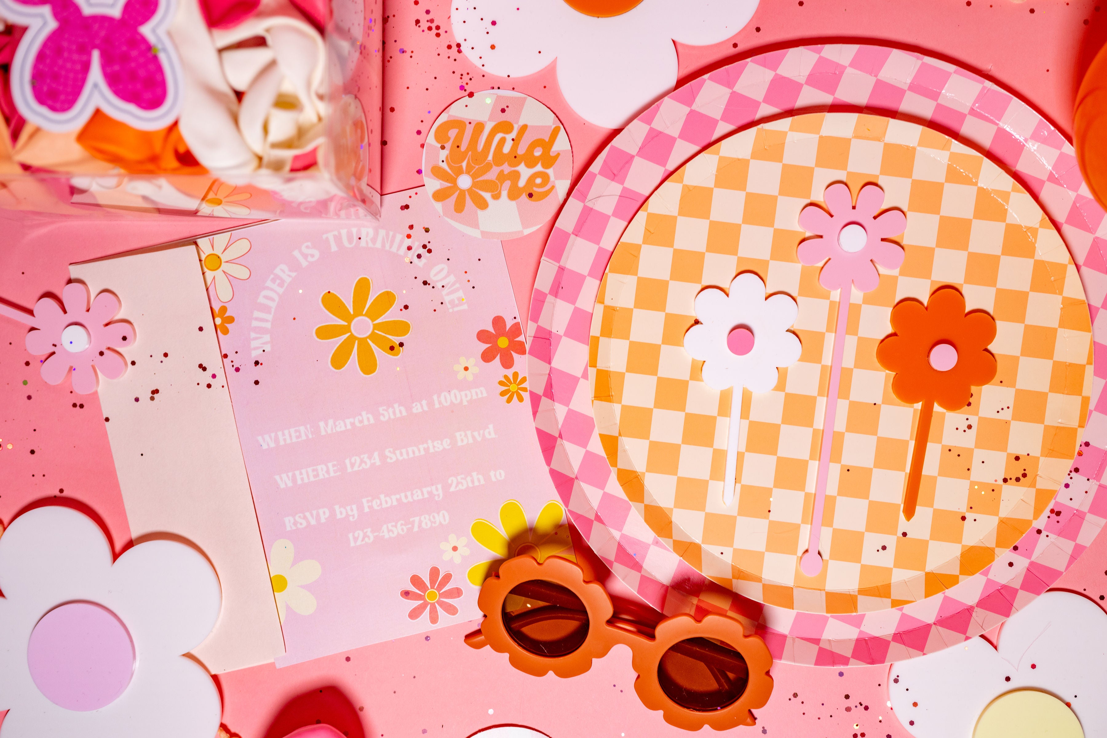 First birthday party inspiration with a Wild One theme, featuring daisy party favors, pink and orange check paper plates, daisy glasses party favors, and a pink and orange balloon arch kit.
