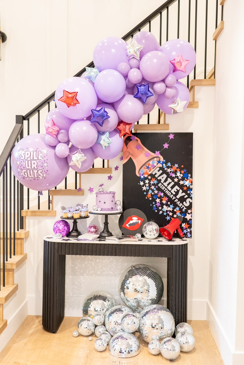 A purple and multicolor metallic star custom balloon garland kit with a custom spill ur guts balloon is used as a decoration idea for an Olivia Rodrigo birthday party featuring cake table decoration ideas.