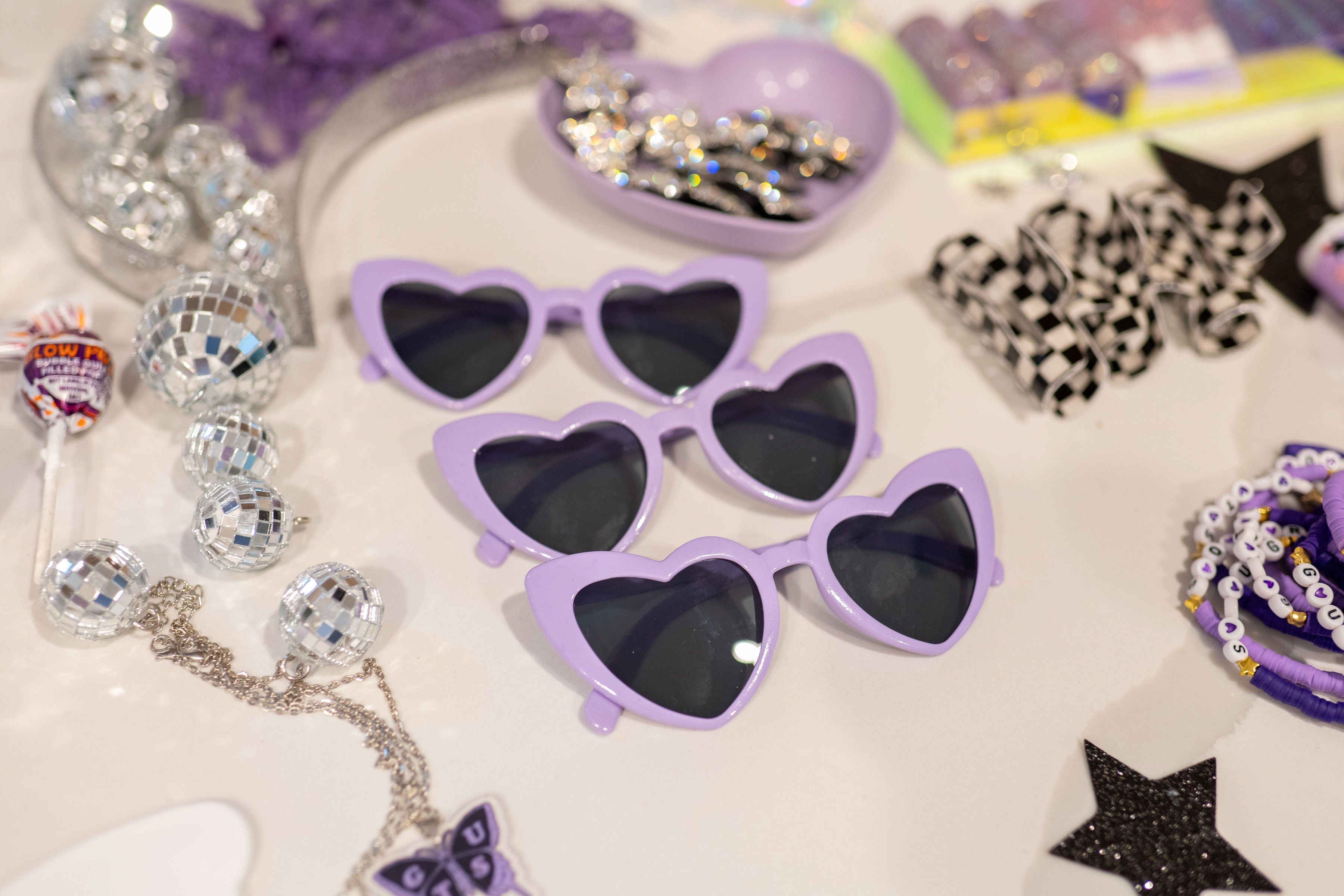 A table filled with Olivia Rodrigo party favors for a tween birthday party, including purple heart-shaped sunglasses, disco ball charms, and a Guts craft kit charm bar.