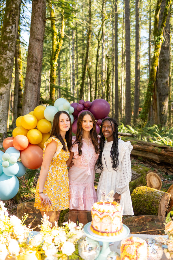 Three girls pose behind a cake table at a Taylor Swift tea party idea for a rustic glam garden party for tweens or teens, or a bridal shower theme idea for Swifties with a multicolor balloon arch kit.