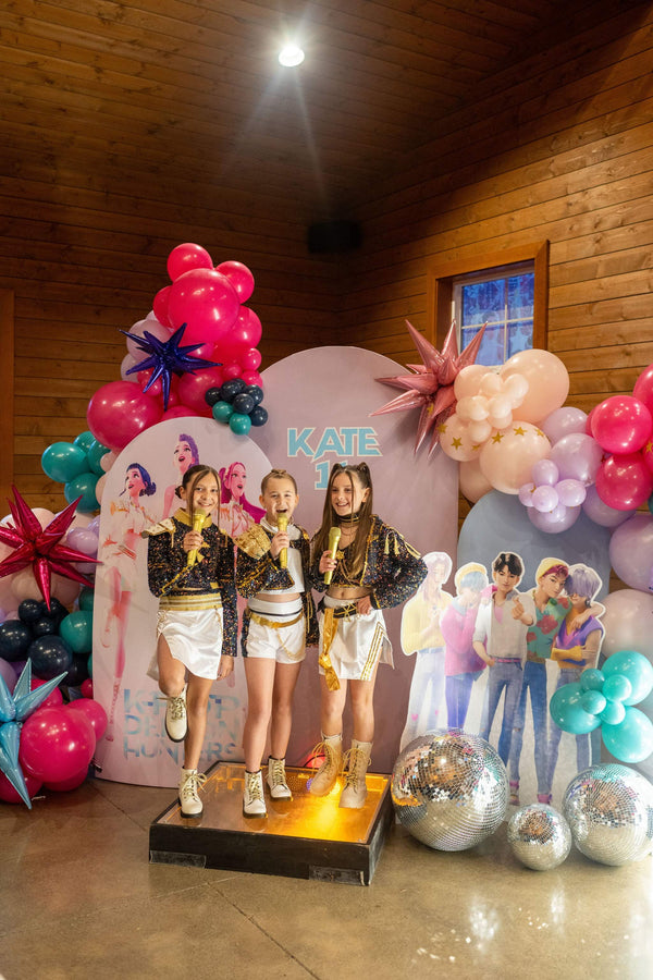 Three girls sing on a stage in front of a  photo shoot backdrop for a kpop demon hunters party for kids or tweens, featuring kpop demon hunters character cutouts and a diy balloon arch kit.