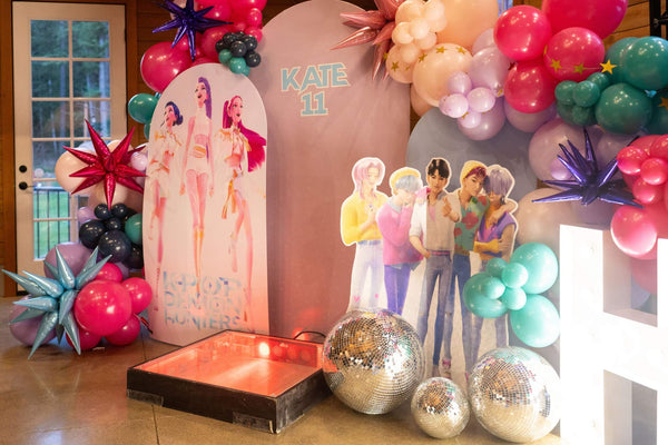A photo shoot backdrop for a kpop demon hunters party for kids or tweens, featuring kpop demon hunters character cutouts and a diy balloon arch kit.